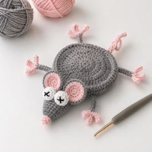 May include: A gray and pink crocheted mouse coaster with a circular body, four limbs with pink paws, and a pointed snout. The mouse features two round ears with pink centers and white eyes with black cross-stitched pupils. A gray yarn ball is nearby.