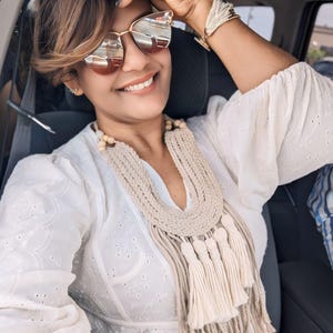 May include: A woman wearing gold-rimmed sunglasses and a white eyelet blouse smiles. She is wearing a macrame necklace with tassels and a woven bracelet. Her nails are painted blue. The woman is sitting in a car.