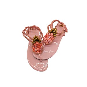 May include: Pink sandals with a thong strap and ankle strap. Each sandal features a pineapple design made of pink and coral-colored stones with a gold-colored crown. The sandals have the signature "Cristina Soriano" on the sole.