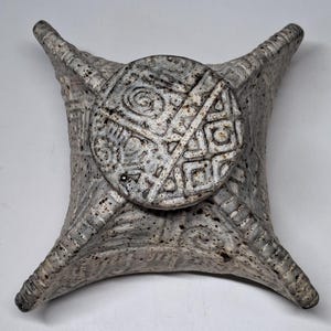 May include: A square-shaped ceramic vessel with a speckled grey finish and four flared corners. The top features a circular lid embossed with geometric patterns, including spirals and cross-hatched lines, which extend down the four sides of the body.