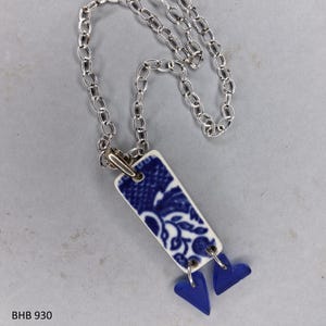 May include: A silver chain necklace with a rectangular pendant featuring a blue and white floral design. Two small blue heart-shaped charms dangle from the bottom of the pendant. The text "BHB 930" is visible.