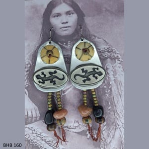 Könnte beinhalten: Ein Paar silberfarbene Ohrringe mit indianischem Design. Jeder Ohrring hat eine kreisförmige Oberseite mit einem braunen und beigen Zentrum, ein Oval mit einem Eidechsenmuster und einen Perlenanhänger mit Erdtönen. Der Text "BHB 160" befindet sich unten.