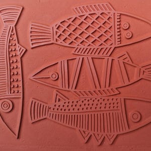 May include: A terracotta-colored rubber stamp featuring a relief design of four fish. One fish is oriented vertically on the left, while three fish are stacked horizontally on the right. Each fish displays distinct geometric patterns and circular eyes.