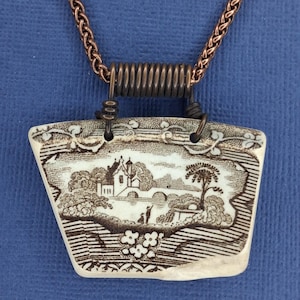 May include: A unique pendant necklace featuring a rectangular ceramic piece with a sepia-toned landscape design. The pendant is suspended from a copper-colored chain and a coiled wire bail. The landscape depicts a building, trees, and figures.