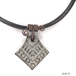 May include: A handcrafted necklace featuring a diamond-shaped pendant with an etched geometric design. The pendant is suspended from a black leather cord and connected with copper wire. The pendant is approximately 1 inch wide.