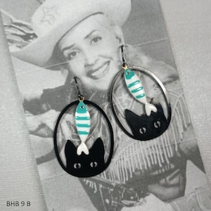 May include: Black cat and fish earrings. The earrings feature a black oval frame with a black cat silhouette at the bottom and a turquoise and white striped fish. The earrings are hanging on a black hook. The text "BHB 9 B" is at the bottom left.