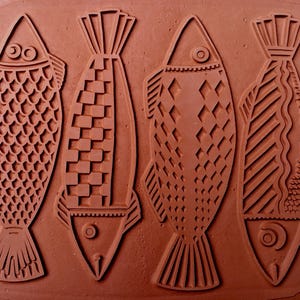 May include: Four stylized fish designs are pressed into a reddish-brown clay surface. Each fish has a unique pattern, including scales, squares, and diamond shapes. The fish tails and fins are also detailed, creating a textured, artistic effect.