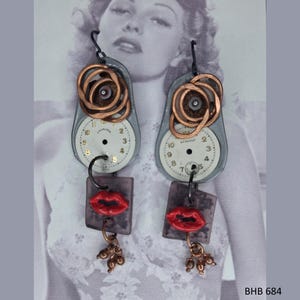 May include: Unique steampunk earrings featuring repurposed watch faces with copper accents. Each earring has a red lip charm and dangling copper beads. The earrings are a creative blend of vintage and industrial design.