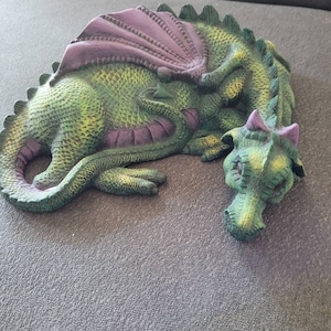 May include: A green and purple sleeping dragon figurine with a textured surface. The dragon has a long tail and a small, curled-up wing.