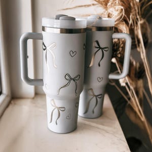 Puede incluir: Dos vasos térmicos grises claros con asas, decorados con lazos y corazones plateados. Los vasos tienen un borde plateado y una tapa blanca. Están sobre una superficie blanca.