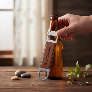 Peut inclure: Une bouteille de bière brune avec un ouvre-bouteille argenté. L'ouvre-bouteille a une poignée en cuir marron avec le texte "Evans Innovations". L'ouvre-bouteille est tenu près du haut de la bouteille. L'arrière-plan est une surface en bois.