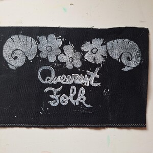 May include: Black fabric patch with white floral and ram's horn designs. The words "Queerest Folk" are written in a stylized font. The patch has a stitched border.