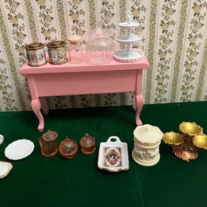May include: A collection of miniature dollhouse accessories including a pink table, two patterned canisters, a clear glass jar, a three-tier white stand, two small cups, three plates, three amber glass jars, a rectangular tray, and a gold-toned stand.