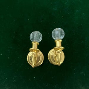May include: A pair of gold-toned metal wall sconce miniatures featuring a circular base, a small arm, and a clear, faceted spherical glass top. Each piece resembles a classic candle holder design with a metallic finish and a transparent orb.