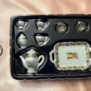 May include: Miniature porcelain tea set with a teapot, cups, saucers, creamer, and a serving tray. The set features floral designs in pastel colors with gold trim. The set is packaged in a black plastic tray.
