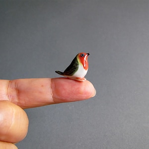 May include: A small ceramic bird figurine with a red breast, white belly, and green and brown wings. The tiny bird is perched on a fingertip against a grey background. The figurine is a detailed representation of a robin.