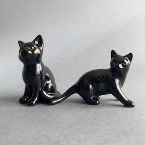 May include: Two glossy black ceramic cat figurines. One cat is in a seated upright position, while the other cat is in a low, prowling stance. Both figurines feature smooth, reflective surfaces and small, alert ears with light-coloured eyes.