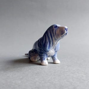 May include: A ceramic figurine of a Basset Hound dog in a seated position, with its head tilted upwards. The dog is primarily blue with white accents on its paws and snout. The figurine has a glossy finish and is set against a gray background.