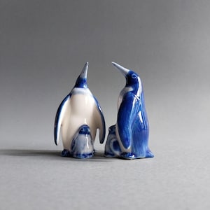 Puede incluir: Un conjunto de figuras de pingüinos de cerámica en tonos azules y blancos. Los pingüinos son de varios tamaños, con dos pingüinos más grandes y dos más pequeños. Las figuras tienen un acabado brillante y están sobre un fondo gris.