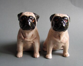 Puede incluir: Dos figuritas de cerámica de carlino, una de color beige y otra marrón, sentadas en posición vertical con ojos y narices negros.