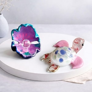 May include: A glass sculpture of a pearl inside a shell and a glass turtle. The shell is painted with shades of blue, purple, and pink. The turtle has pink flippers and a white shell with blue and green spots.