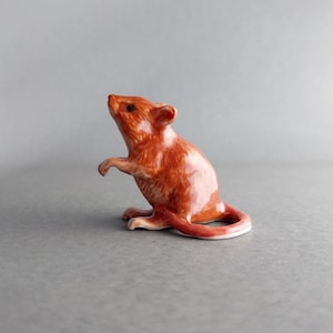 May include: A ceramic figurine of a mouse in a standing pose. The mouse is primarily orange with a glossy finish, and its tail curves around. The figurine has small, detailed ears and paws, and is set against a gray background.