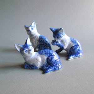 以下が含まれることがあります： 青と白のセラミック製の猫の置物3つ。猫は様々なポーズをとっており、横たわっているもの、座っているもの、身繕いをしているものがあります。置物は光沢のある仕上げで、装飾品として使用される可能性があります。