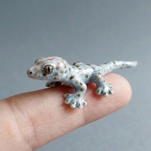 May include: A small ceramic gecko figurine. The gecko is pale blue with brown spots and a white head. The figurine is resting on a finger, highlighting its small size and detailed design.