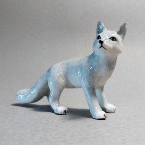 Peut inclure: Figurine en céramique représentant un renard en position debout, la tête levée. Le renard est de couleur bleu clair et blanc, avec des accents noirs sur les yeux et les pattes. Objet décoratif.