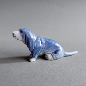 May include: A ceramic figurine of a Basset Hound dog in a seated position. The dog is primarily light blue with white accents on the face, paws, and tail tip. The figurine has a glossy finish and is set against a gray background.