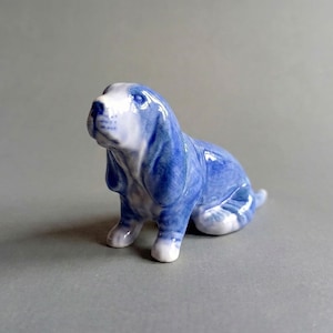May include: A small ceramic figurine of a dog with long, drooping ears, featuring a glossy white and blue marbled glaze finish. The dog is depicted in a seated position with its head tilted slightly upward, showcasing detailed facial features.