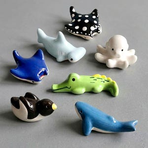Peut inclure: Un ensemble de sept figurines d'animaux en céramique comprenant une raie noire et blanche à pois, un requin-marteau bleu clair, une pieuvre blanche, une raie bleue unie, un crocodile vert, un pingouin noir et blanc et une figurine de baleine bleue.