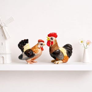 May include: Two ceramic rooster figurines. One rooster is standing, the other is sitting. Both have red combs, yellow beaks, and black, brown, and yellow feathers. They are on a white shelf with a white windmill and a small vase of flowers.