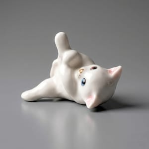 May include: A small white ceramic kitten figurine lying on its side with one paw raised. The cat has bright blue eyes, a small pink nose, and delicate pink-tinted ears, with a glossy finish that reflects light across its smooth, curved body.