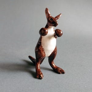 Op de afbeelding: Een bruin en wit keramisch kangoeroebeeldje. De kangoeroe staat rechtop met de armen omhoog en de oren omhoog. Het beeldje heeft een glanzende afwerking en is een decoratief item.