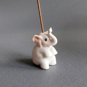 May include: A white ceramic elephant incense holder with a glossy finish, featuring pink-tinted ears and cheeks, a raised trunk, and a small hole on the top of its head holding a single thin brown wooden incense stick in an upright position.