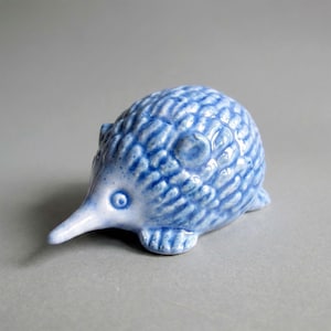 May include: A small ceramic figurine of an echidna with a glossy finish. The sculpture features a light blue and white mottled glaze, a pointed snout, a rounded body with textured spines, and small feet, capturing the animal's distinct form.