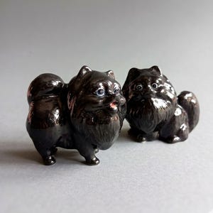 Könnte beinhalten: Zwei kleine, glänzend schwarze Keramikfiguren in Form von Zwergspitz-Hunden. Jede Figur weist eine detaillierte Fellstruktur, spitze Ohren, einen geringelten Schwanz und bemalte Augen auf. Der linke Hund hat ein leicht geöffnetes Maul.