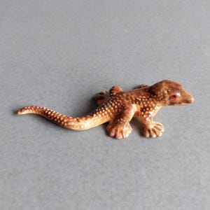Op de afbeelding: Een kleine keramische hagedissenfiguur in bruin- en beigetinten met witte stippen. De hagedis heeft rode ogen en ligt op een grijze ondergrond. Het beeldje is een decoratief item.