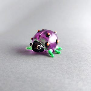 May include: A small, glass ladybug figurine. The ladybug has a purple body with orange and black spots, black head, and green legs. The figurine is a decorative item.