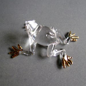 以下が含まれることがあります： 金色の足を持つ透明なガラス製のカエルの置物。 カエルは這うような姿勢で、手足が伸びています。 この置物は細部までこだわって作られており、手作りのようです。