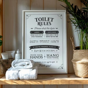 Könnte beinhalten: Ein gerahmtes schwarz-weißes Schild mit dem Text "TOILET RULES" und Badezimmeranweisungen. Darunter gefaltete weiße Handtücher, ein Korb mit Toilettenartikeln und eine Topfpflanze.