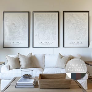 May include: Three framed topographic map prints with black frames, each labeled with a mountain name: Ben Nevis, Scafell Pike, and Yr Wyddfa. The prints are displayed above a white sofa with beige pillows. A wooden coffee table holds a woven basket and books.