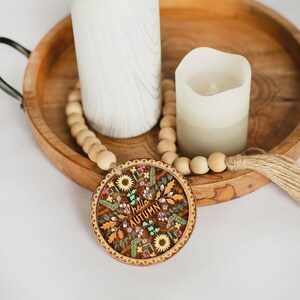 Puede incluir: Una bandeja de madera con un círculo decorativo de madera con las palabras "hello AUTUMN" rodeadas de un diseño floral. Dos velas blancas y una guirnalda de cuentas también están en la bandeja.