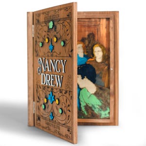 May include: Wooden book-shaped box with the title "NANCY DREW" in white lettering. The cover is intricately carved with floral designs and colorful accents. The open box reveals a painted scene of figures.
