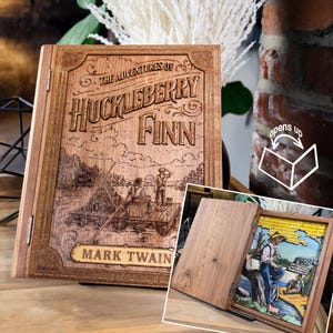 Op de afbeelding: Een houten doos in de vorm van een boek met een scharnierend deksel, lasergegraveerd met de tekst The Adventures of Huckleberry Finn door Mark Twain en een illustratie van twee figuren op een vlot. De binnenkant toont een kleurrijke 3D-scène.