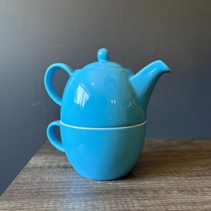 May include: A vibrant turquoise ceramic tea set, comprising a teapot with a lid and a cup. The teapot rests on the cup, forming a neat stack. The set is a solid, glossy turquoise, ideal for serving tea. A charming addition to any kitchen.