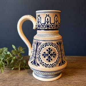 May include: A ceramic beer stein featuring a beige base with intricate cobalt blue relief patterns. The design includes a central cross motif surrounded by ornate scrollwork, a sturdy curved handle, and a textured rim with a repeating geometric border.