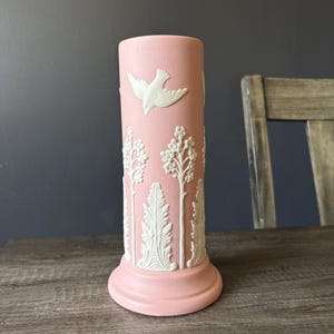 May include: A pink ceramic vase with white relief detailing. The vase features a flying dove and floral and leaf patterns. The vase has a cylindrical shape with a flared base.