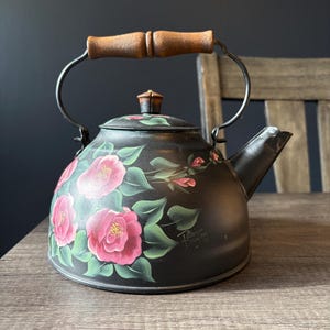 Peut inclure: Une bouilloire à thé en métal noir ornée de fleurs de camélia roses peintes à la main avec des feuilles vertes. La bouilloire possède un bec verseur incurvé, un couvercle avec un petit bouton en bois et une anse articulée avec une poignée en bois brun texturé. La signature Patricia Day est visible.